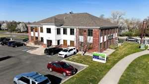 A two story brick and white exterior office building at 16205 W 64th Ave in Arvada with parked cars in the lot, landscaped grass areas, blooming trees, and clear blue sky.
